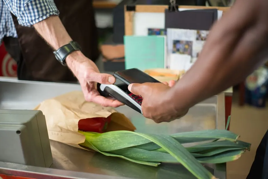 A shopper paying with a smartphone at a grocery store checkout counter next to fresh produce